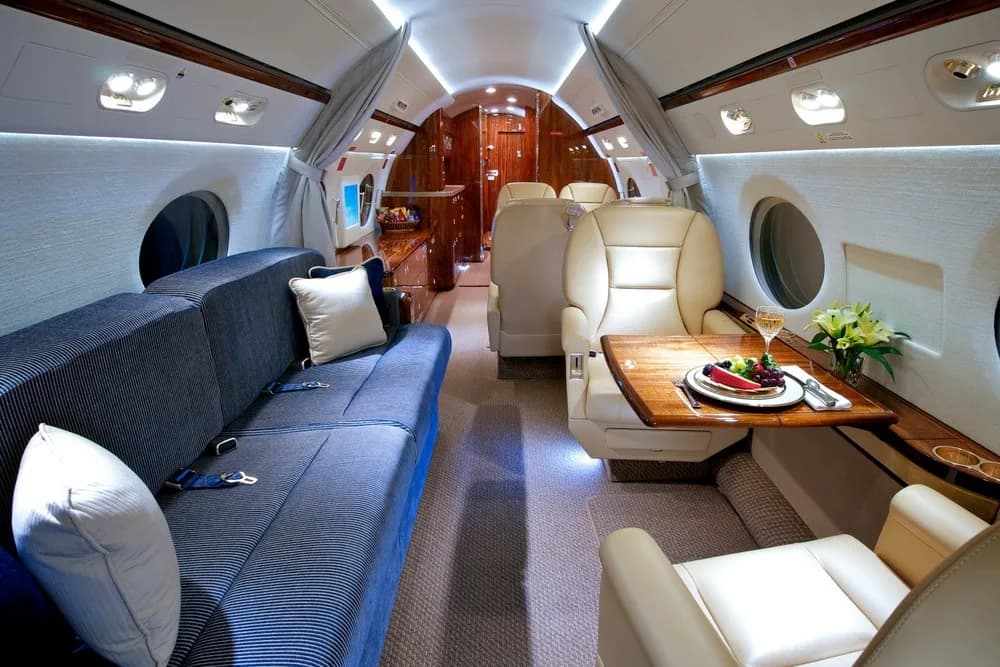 The luxurious cabin interior of a private jet, showing a blue striped sofa, tan leather seats, and a wooden table with a meal.