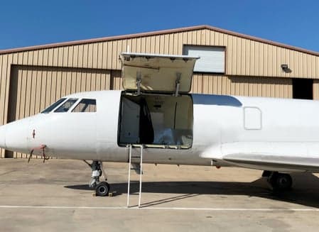 Side view of an aircraft with an open door and a ladder, parked in front of a hangar.
