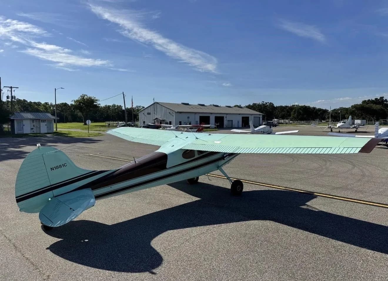 A rear-side view of a brown and light green Cessna 170B (N1881C) parked on an airport apron with hangars in the background.