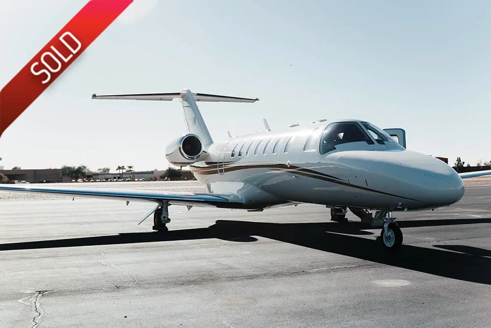 A white business jet with a brown stripe is parked on an asphalt tarmac under a clear sky.