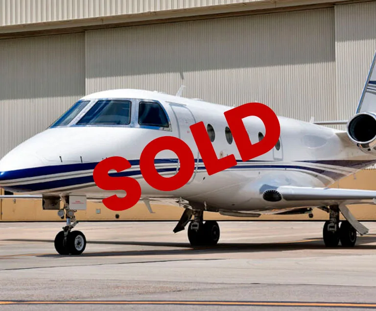 A white business jet with blue stripes is parked on a tarmac in front of a hangar, with the word 'SOLD' overlaid in red.