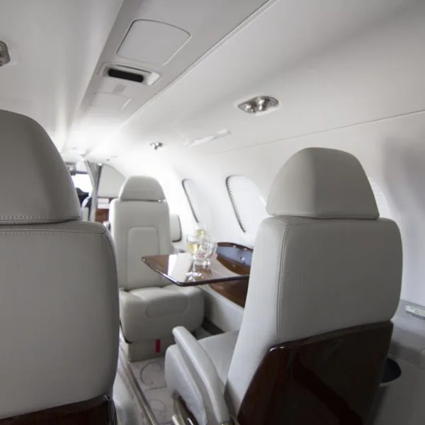 A side view of the private jet's interior, highlighting light grey leather seats and a small wooden table with drinks.