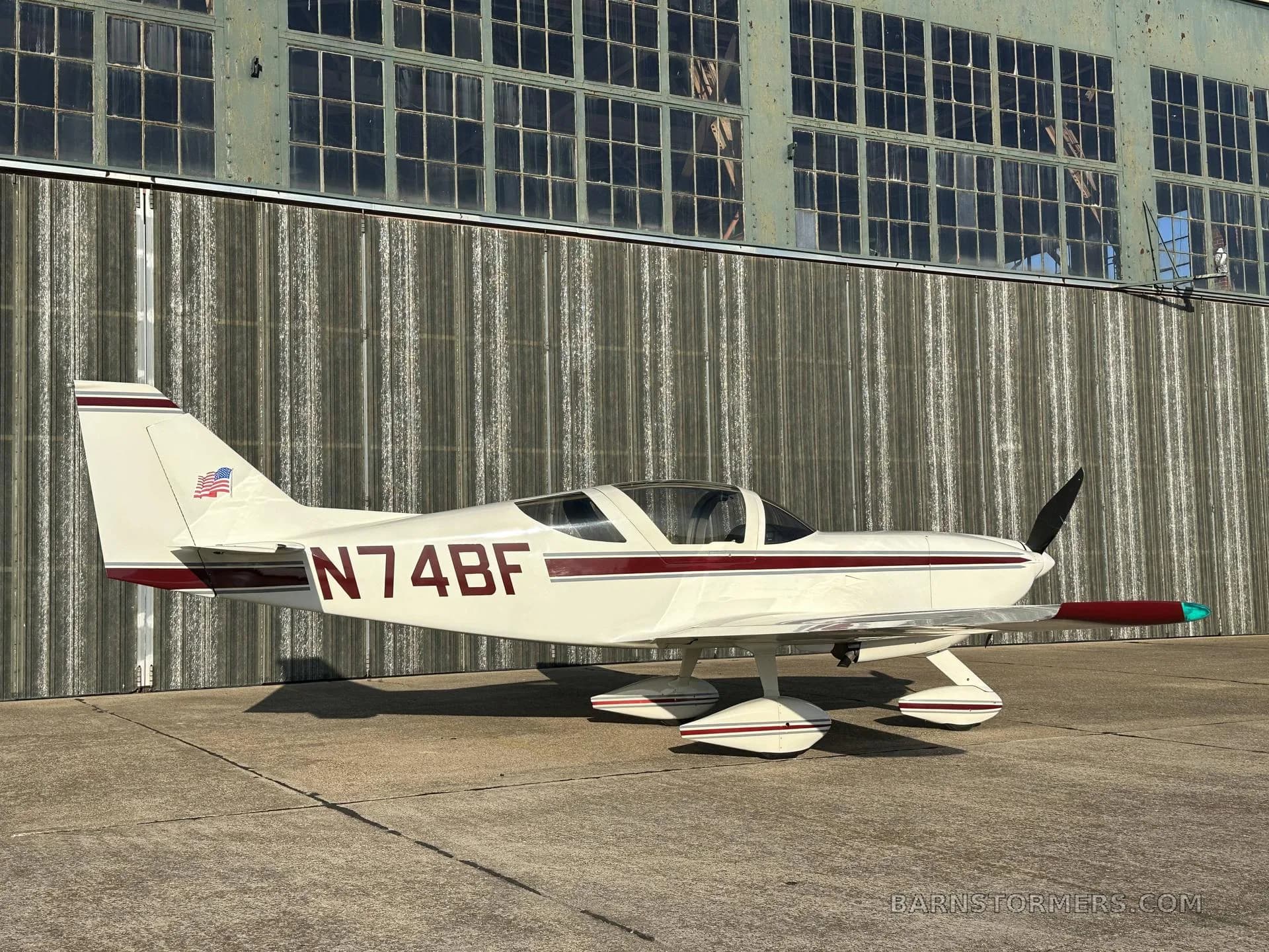 A white single-engine aircraft with a maroon stripe and registration N74BF is parked on concrete, viewed from the left side, in front of a hangar.