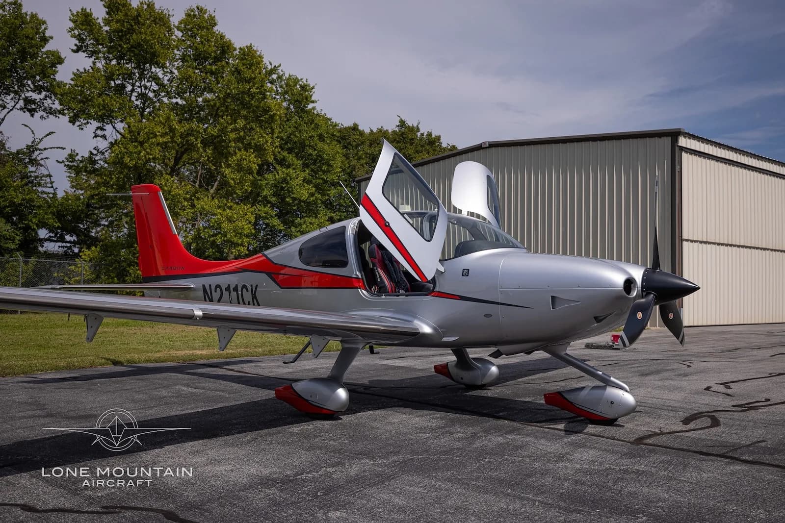 A low-angle front-side view of a silver and red Cirrus SR22T aircraft with registration N211CK parked on an asphalt ramp with its gull-wing doors open.