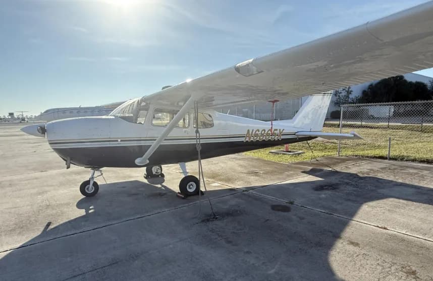A side view of a white and black Cessna 172 with registration N6865X, parked on an airfield under bright sunlight.