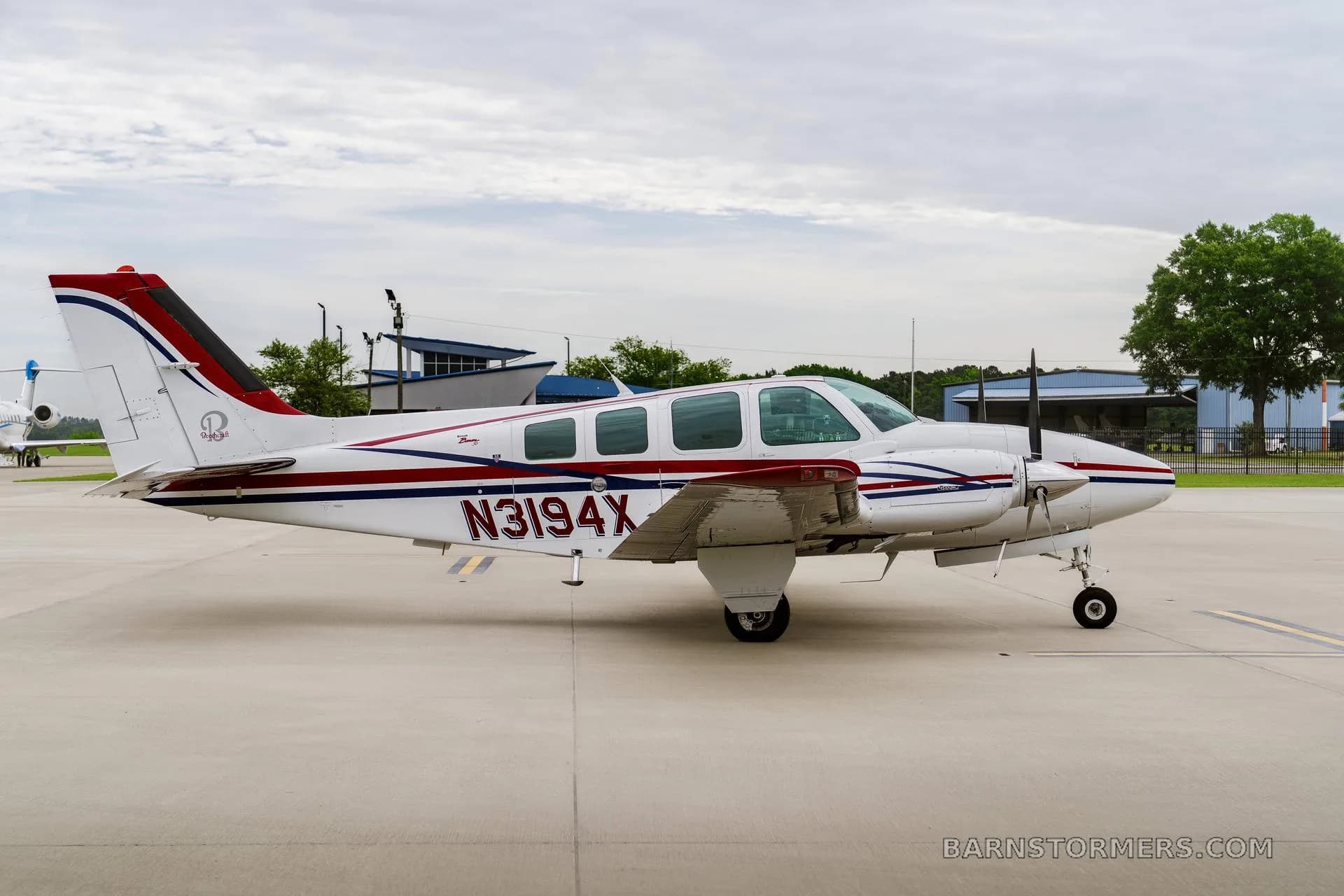 A side view of a white twin-engine propeller aircraft with red and blue stripes, registration N3194X, parked on an airfield with buildings in the background.