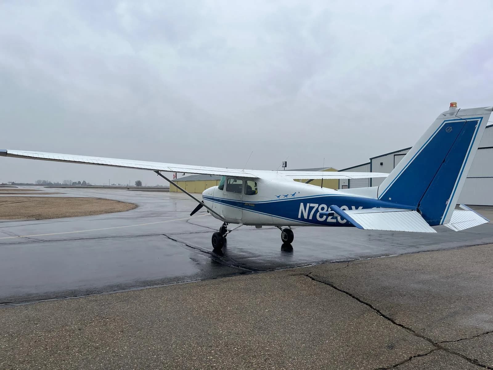 A rear-side view of a white Cessna 172 Skyhawk with blue stripes and tail, registration N7820X, parked on a wet tarmac.