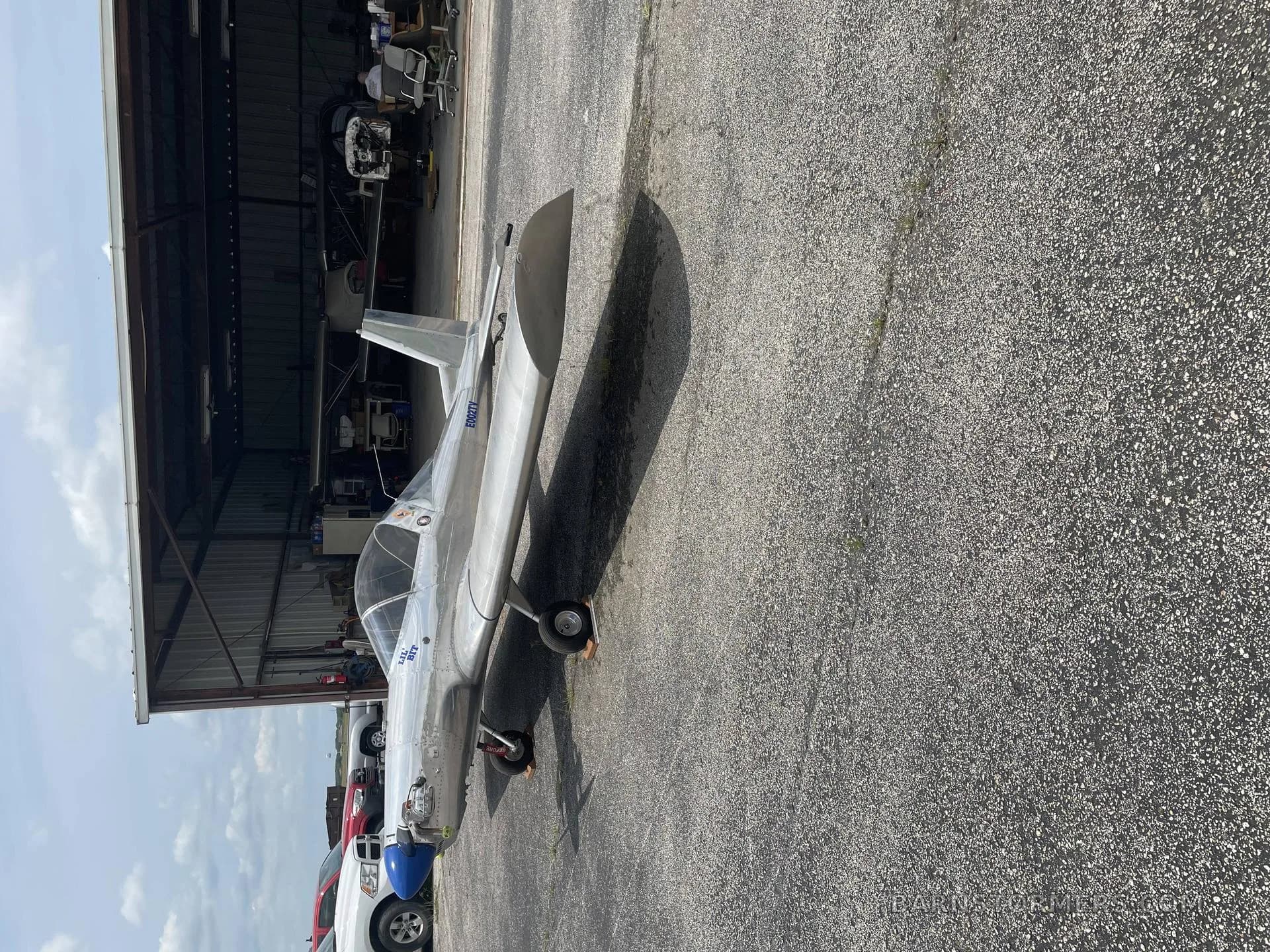 A silver single-engine aircraft with a blue nose cone is parked on an asphalt apron next to an open hangar.