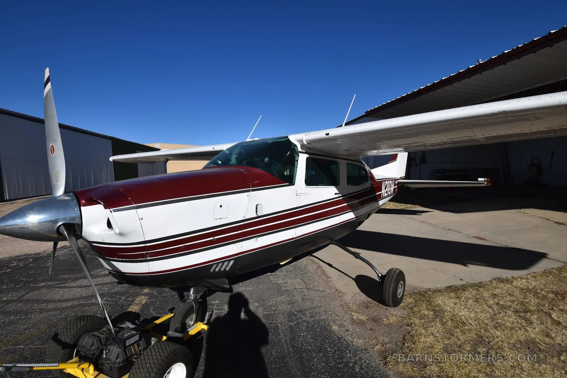 A close-up side view of a white and maroon Cessna 210 aircraft with registration N210W, parked on asphalt next to a hangar.