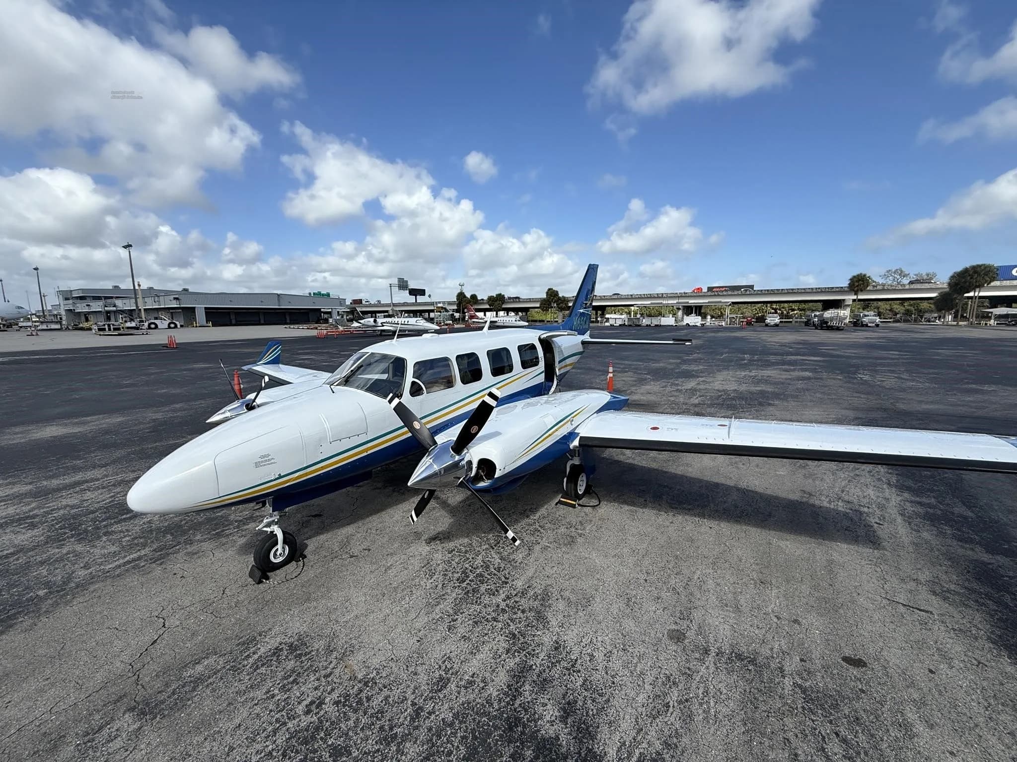 A white and blue twin-engine propeller aircraft with yellow and green stripes, parked on an airport tarmac with its passenger door open.