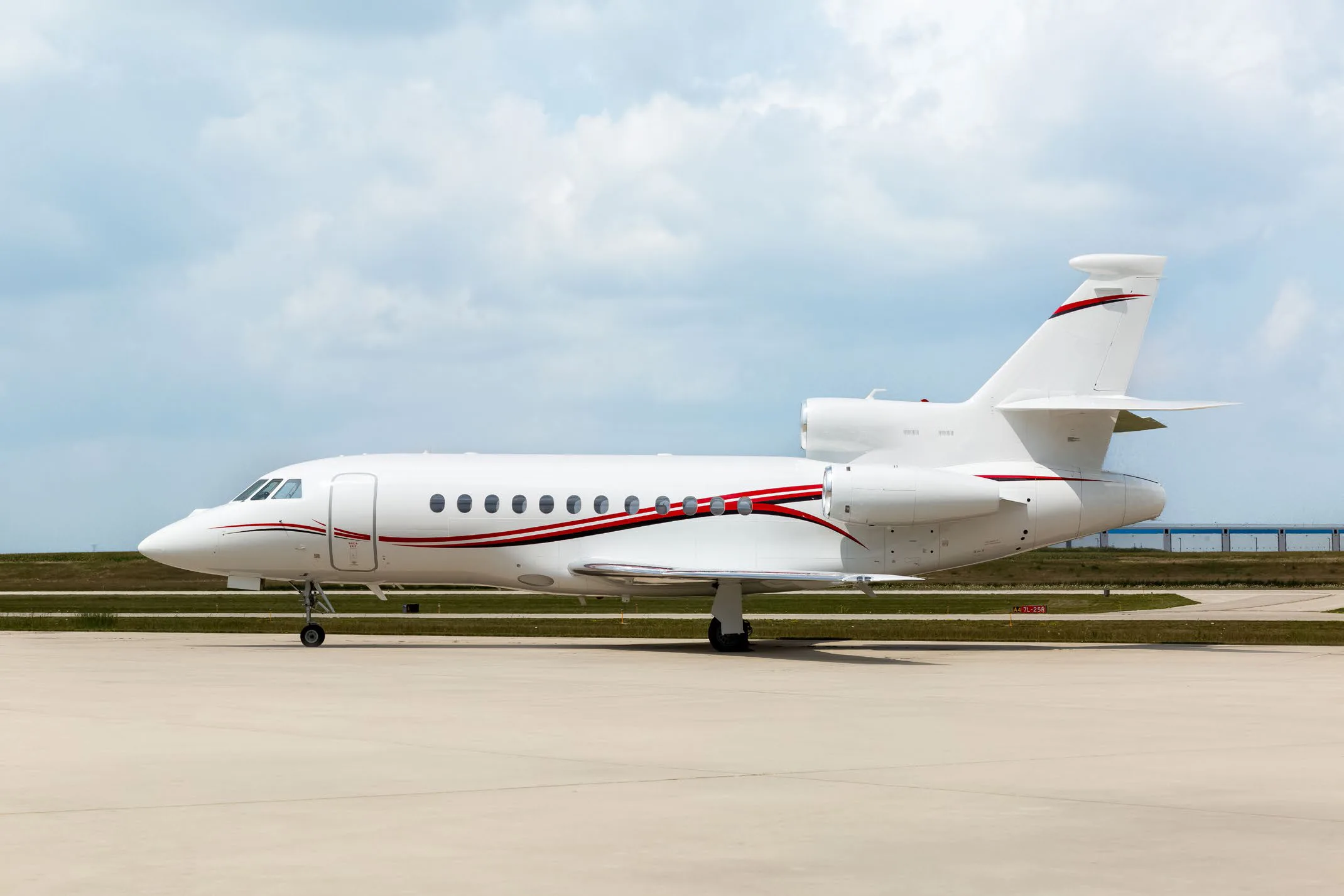 A white Dassault Falcon business jet with red and black stripes on the fuselage and tail, parked on an airport tarmac under a cloudy sky.