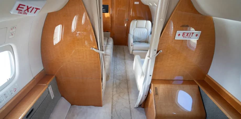 A view down the aisle of a private jet cabin, showing light wood paneling and "EXIT" signs above the doors.