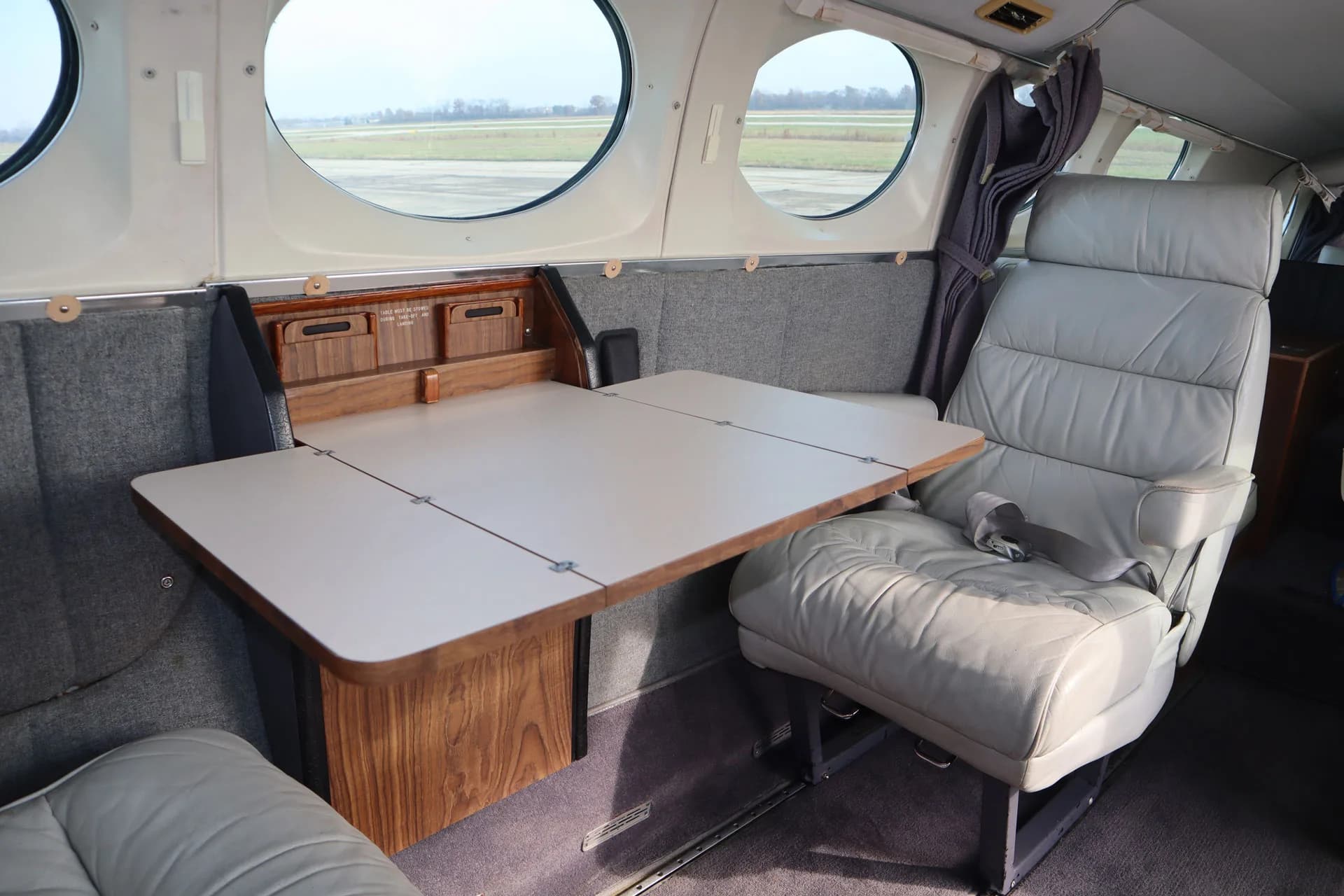 The interior of the aircraft features a table and gray leather seat with a window in the background.