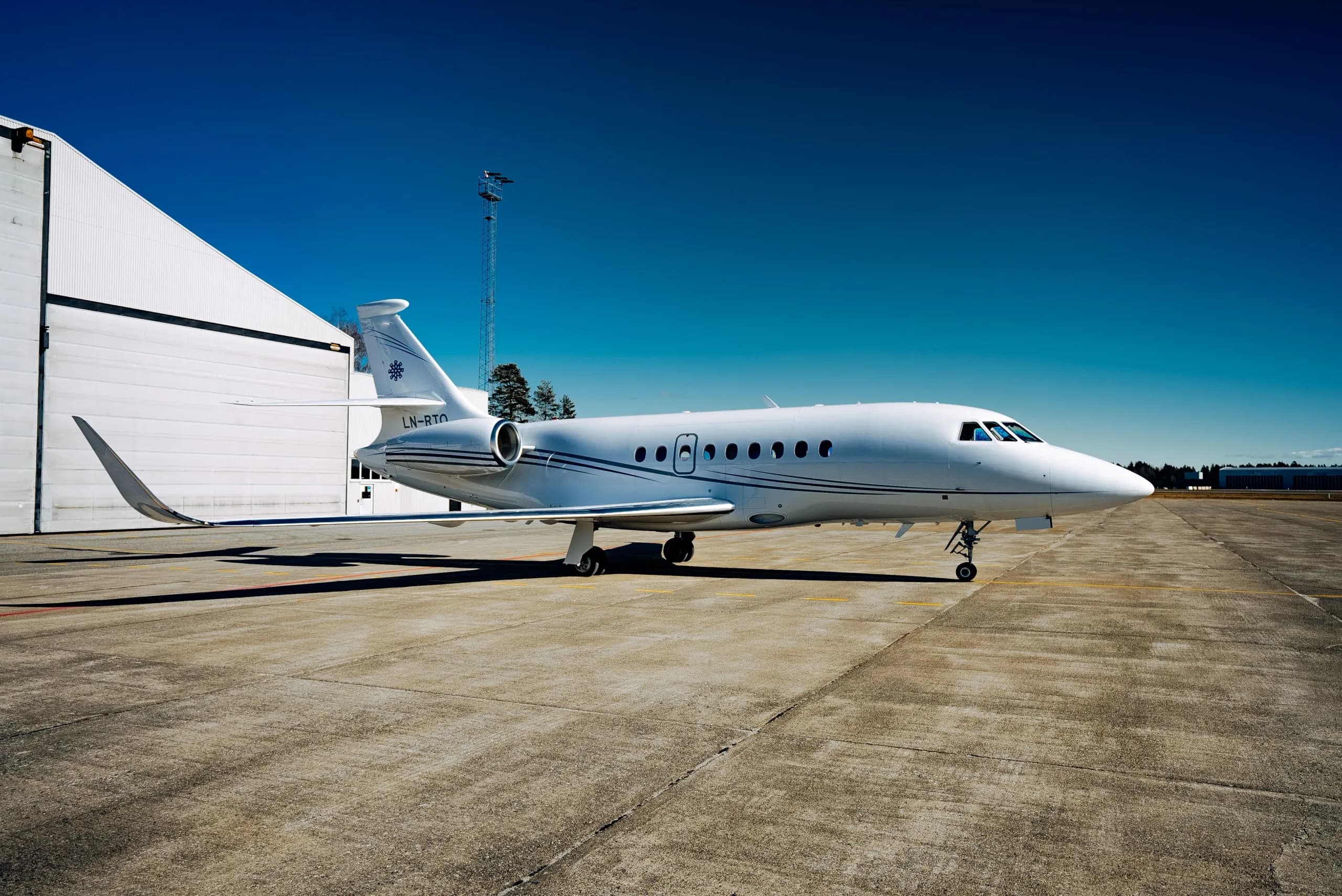 A white Dassault Falcon business jet with blue stripes and registration LN-RTO parked on a tarmac next to a white hangar.