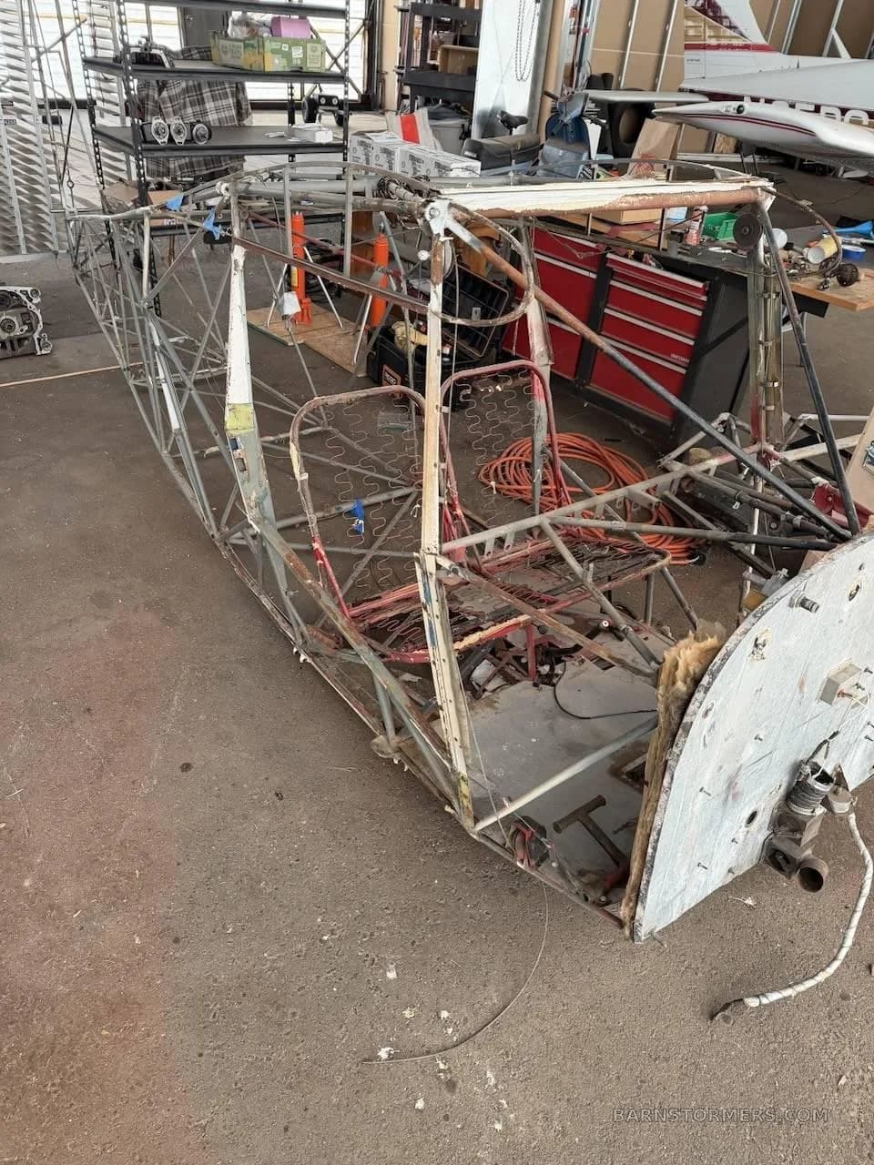 A stripped-down aircraft fuselage frame with exposed internal structure and seat frames in a hangar.