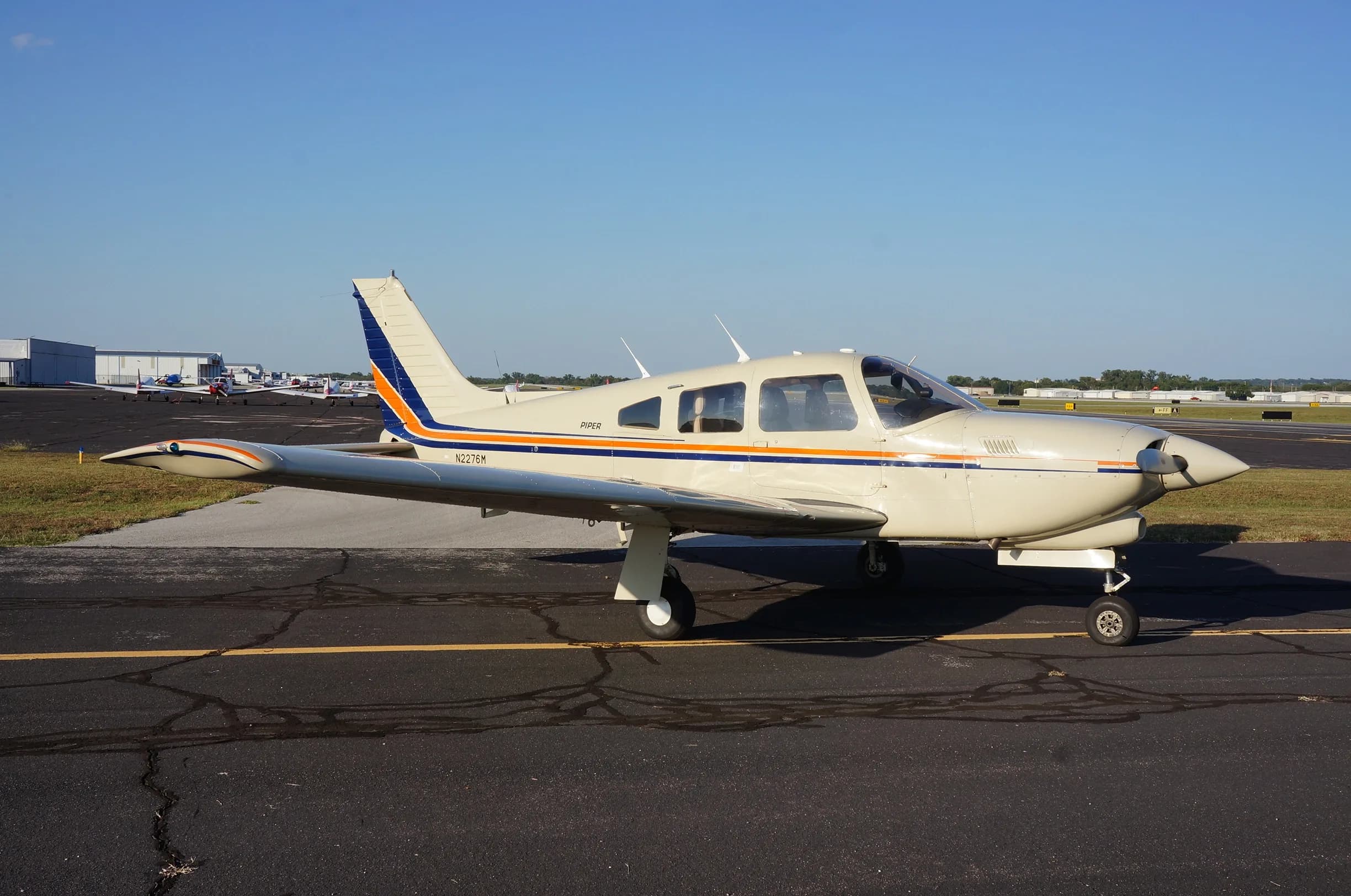 A cream-colored Piper aircraft, registration N2276M, with blue and orange stripes, parked on an asphalt tarmac.
