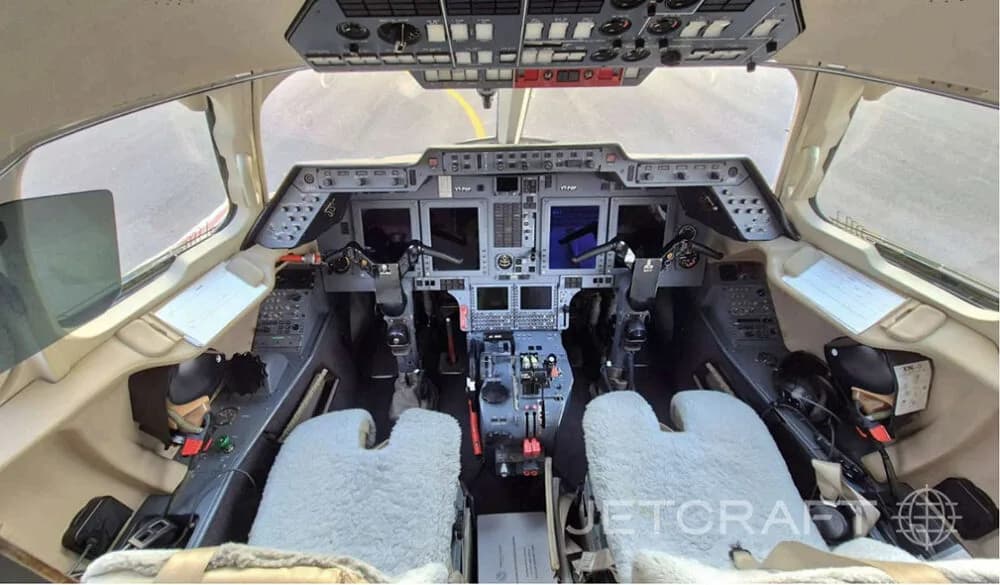 The cockpit of a business jet features multiple display screens and two pilot seats with furry covers.