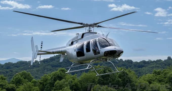 A white Bell 206 JetRanger helicopter with registration N110JJ flying over green treetops under a blue sky.