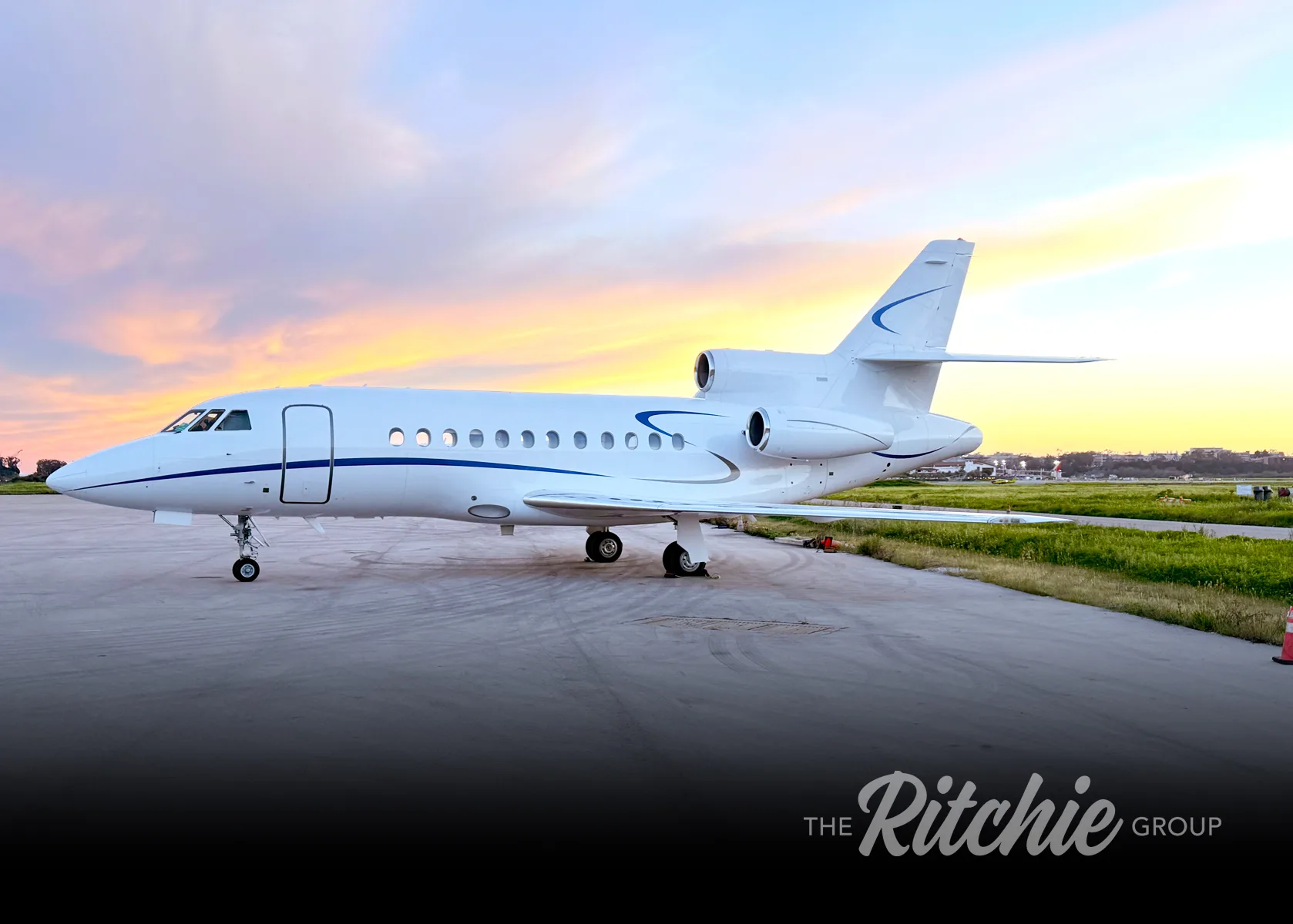A white business jet with blue accents on the tail and fuselage, parked on an airfield at sunset.