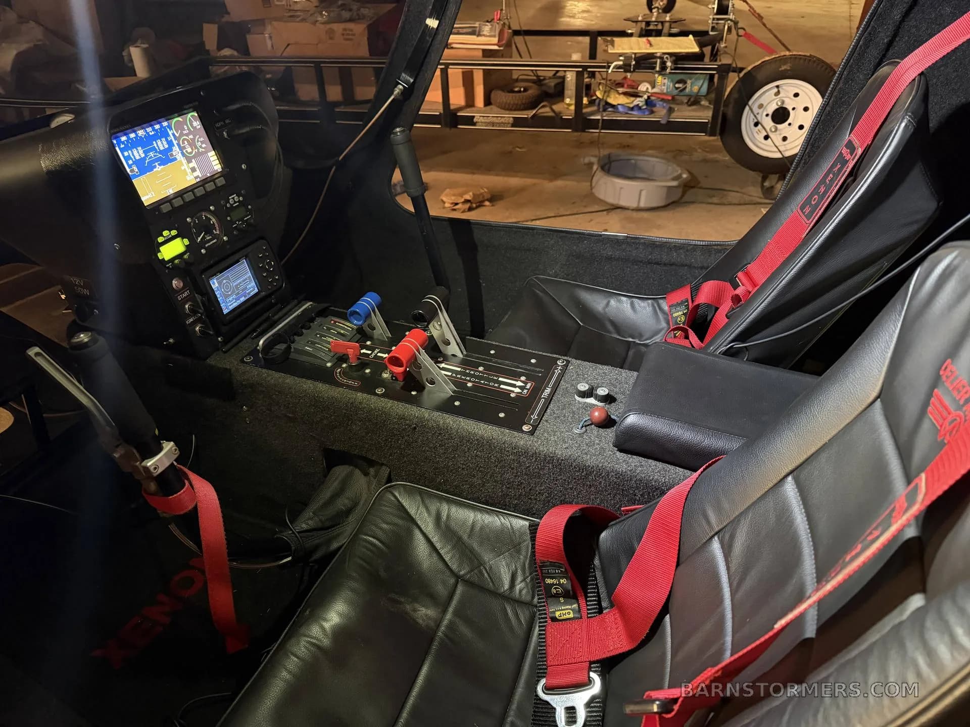 The cockpit of a red autogyro features a digital instrument panel, flight controls, and two black leather seats with red harnesses.