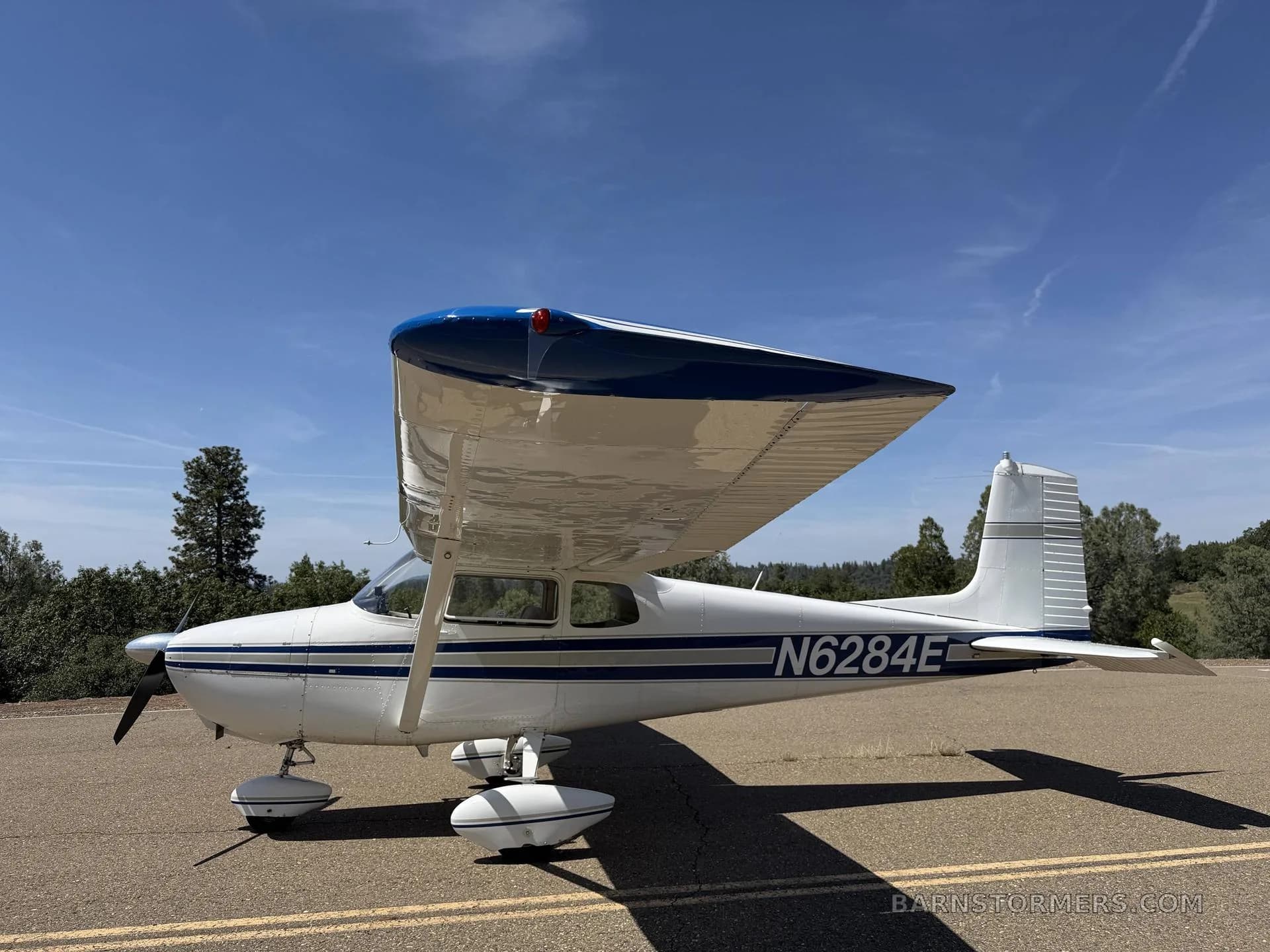 A white and blue single-engine high-wing aircraft, N6284E, parked on an asphalt strip under a clear blue sky.