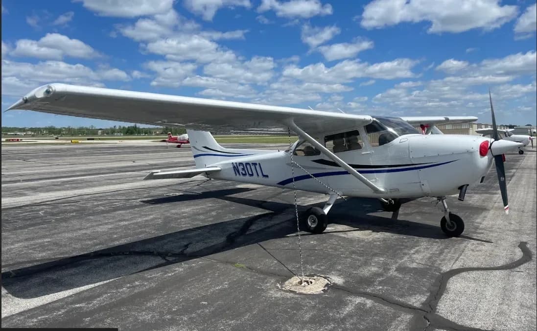 A white single-engine Cessna aircraft, N30TL, with blue stripes parked on an apron under a cloudy sky.