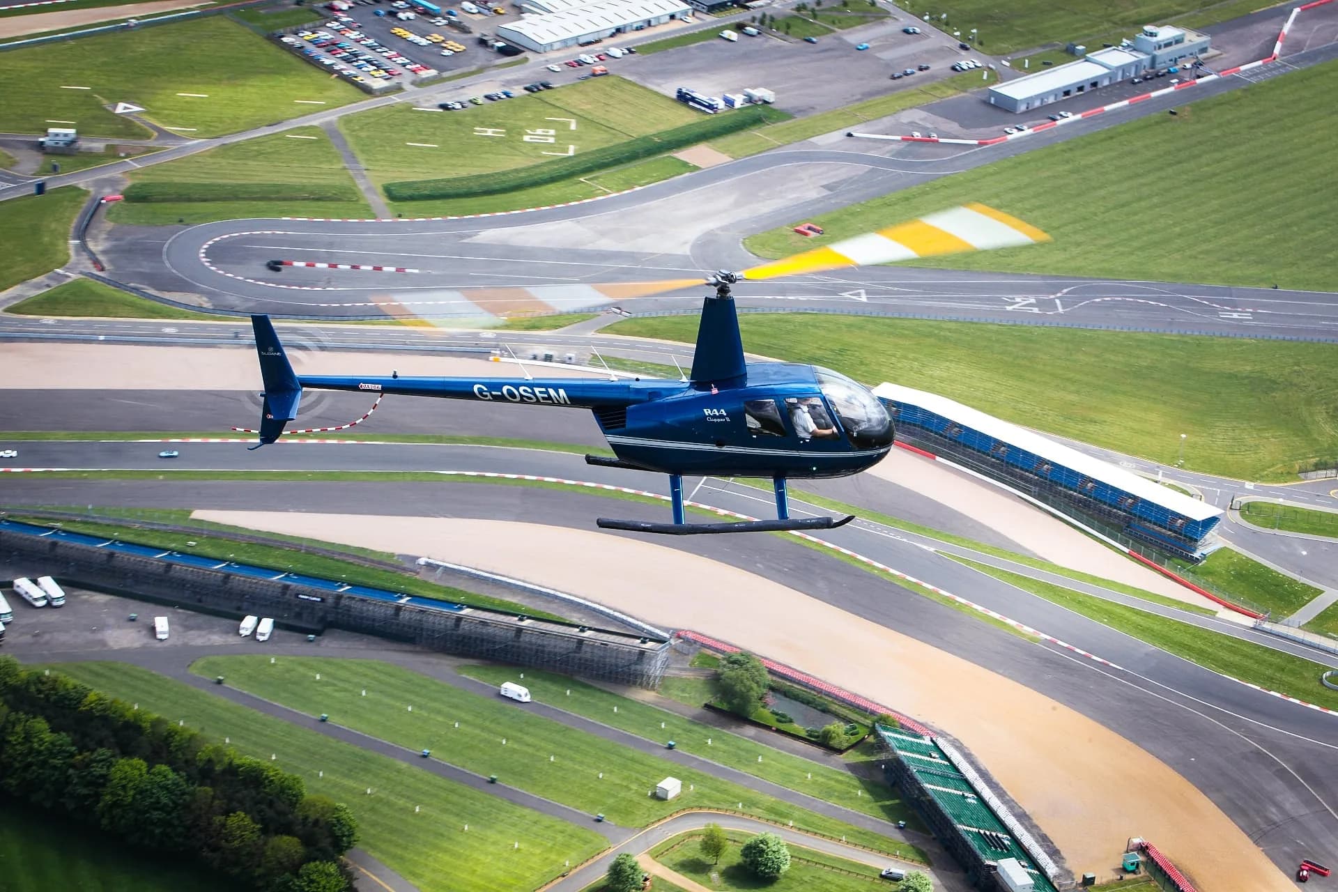 A blue Robinson R44 helicopter flying over a racetrack and parking lot.