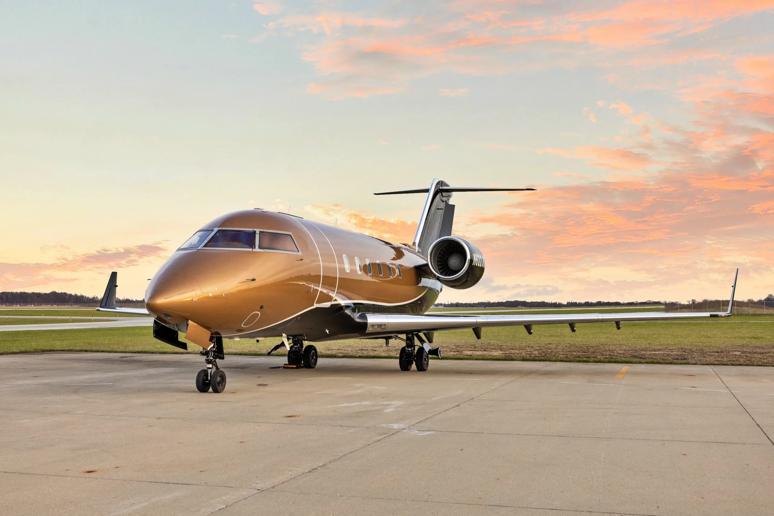 A gold and black Bombardier Challenger private jet is parked on an airport tarmac under a sunset sky.