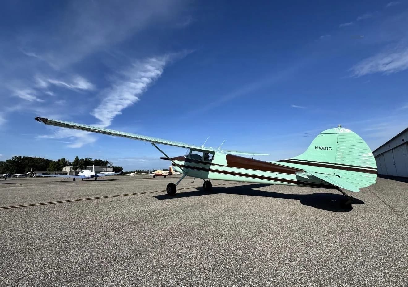 A rear-side view of a brown and light green Cessna 170B (N1881C) parked on an airport tarmac under a blue sky.