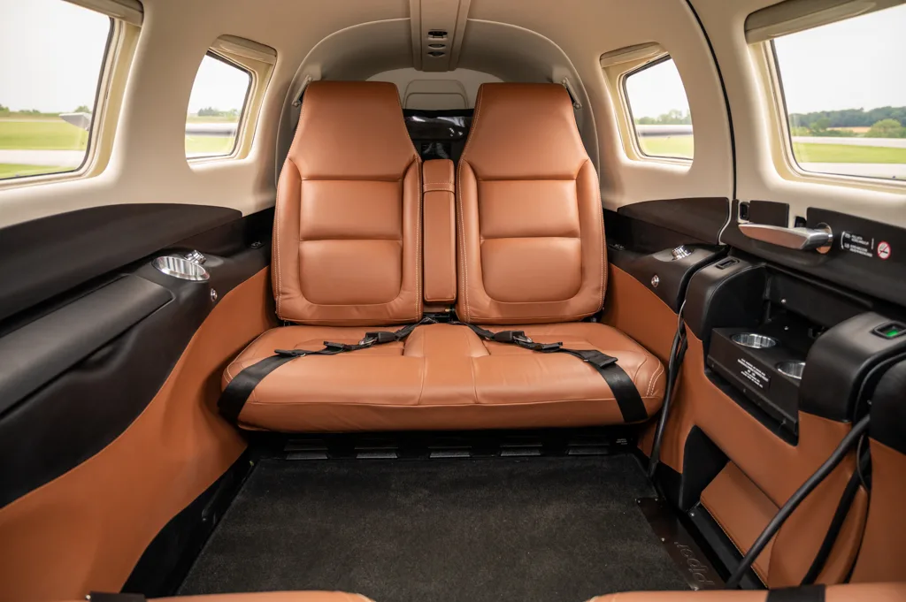 The rear cabin interior of an aircraft, featuring two tan leather seats with black seatbelts and side panels.