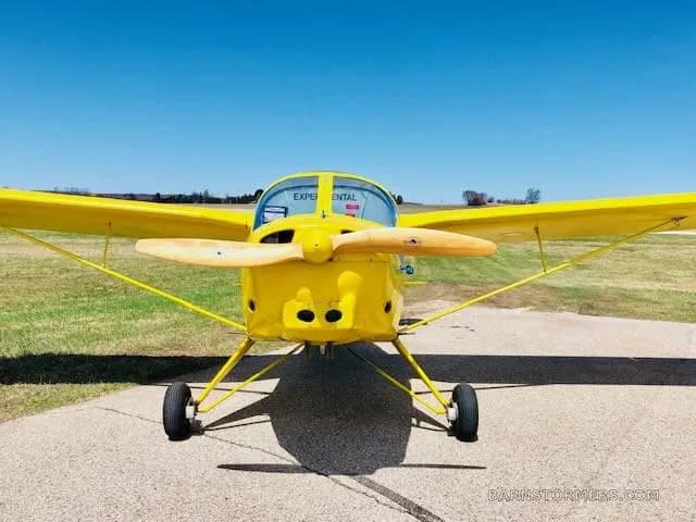 A bright yellow single-engine experimental aircraft with a wooden propeller, viewed head-on, parked on an asphalt surface.