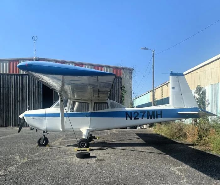 A white and blue high-wing aircraft, registration N27MH, is parked on an asphalt apron with hangars in the background.