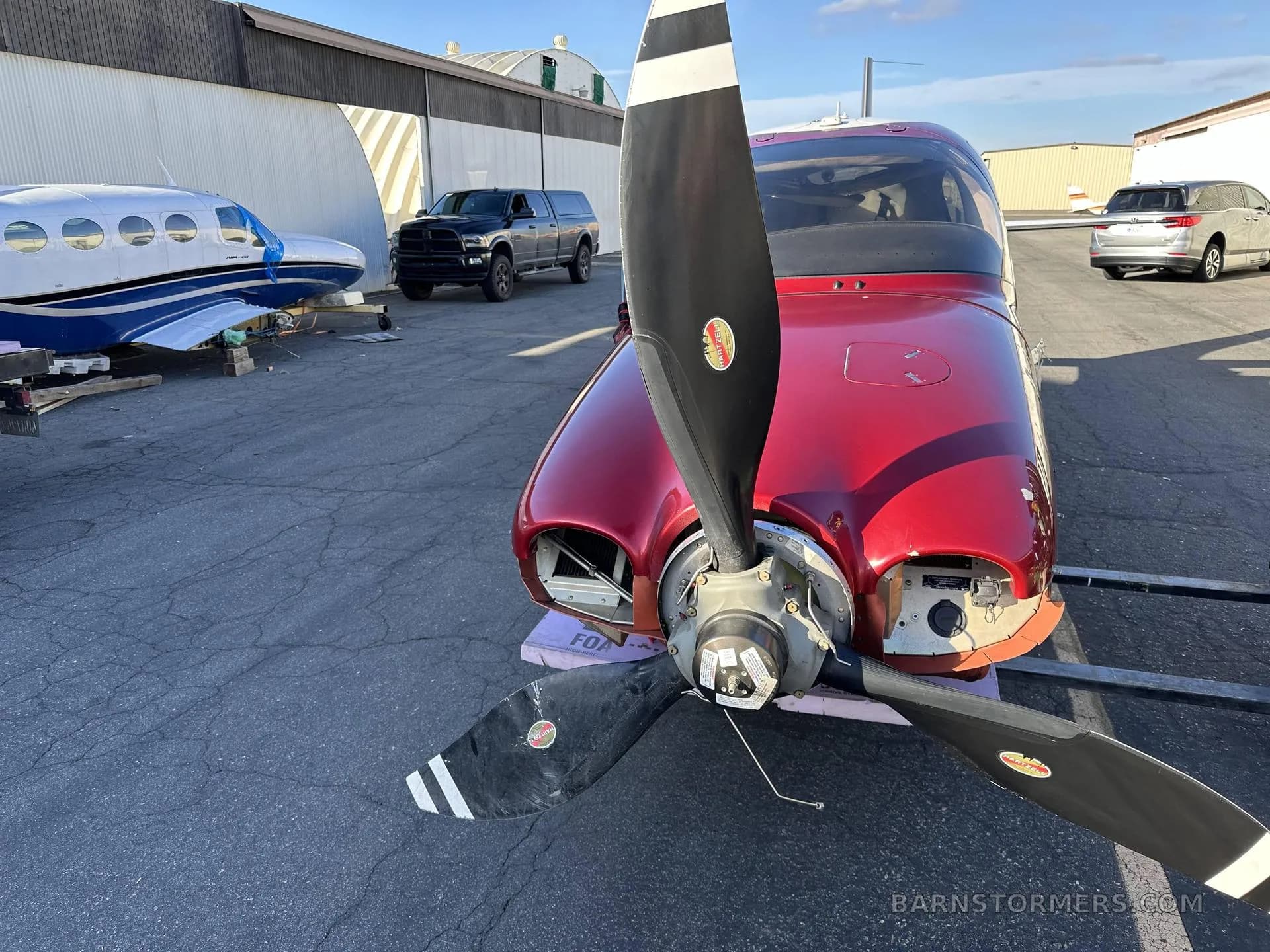 A red single-engine aircraft with its cowling removed, showing the propeller, parked outdoors with other vehicles and aircraft in the background.