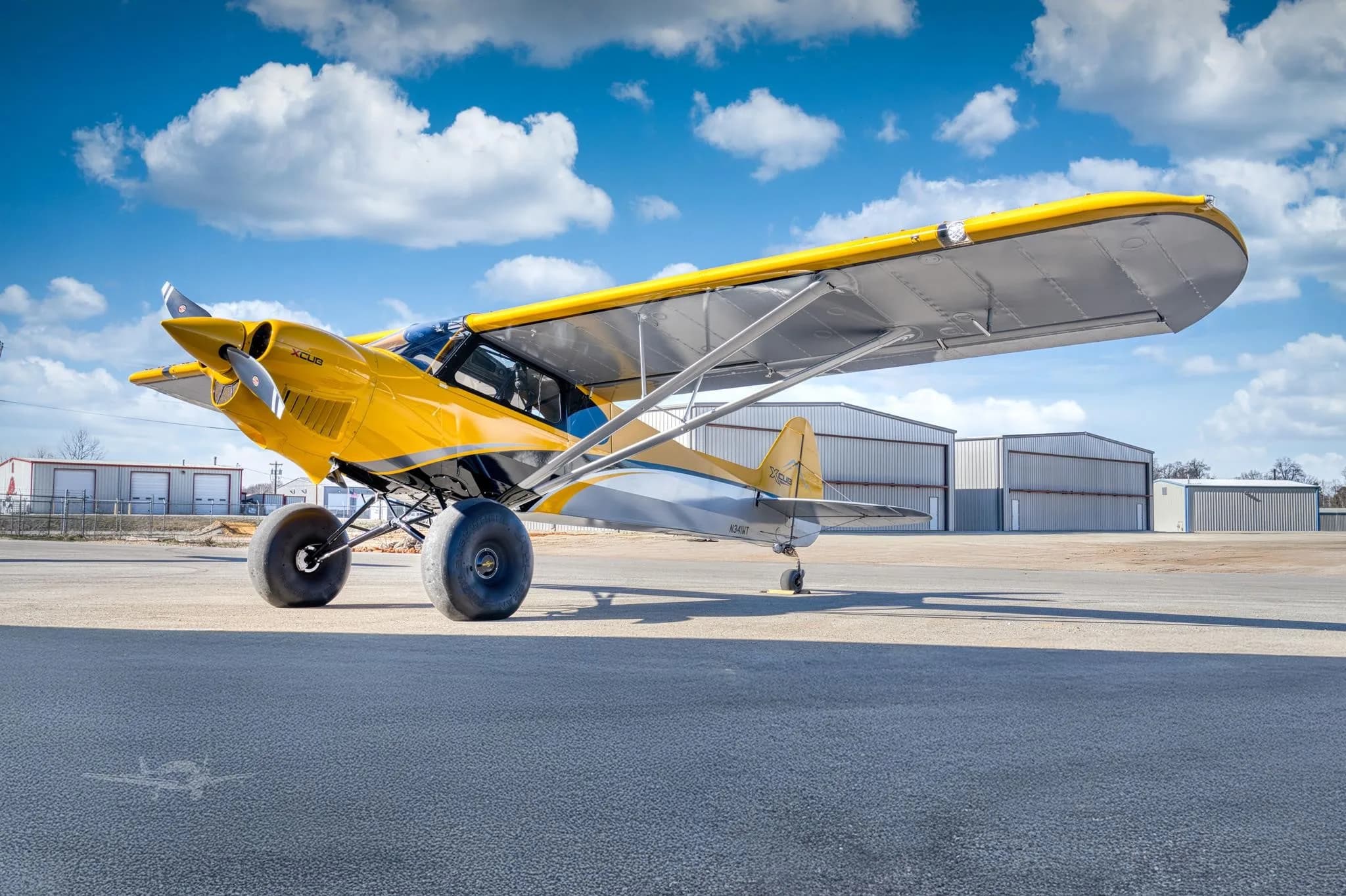 A yellow and grey X-Cub aircraft with large tundra tires is parked on an airfield apron with hangars in the background under a blue sky.