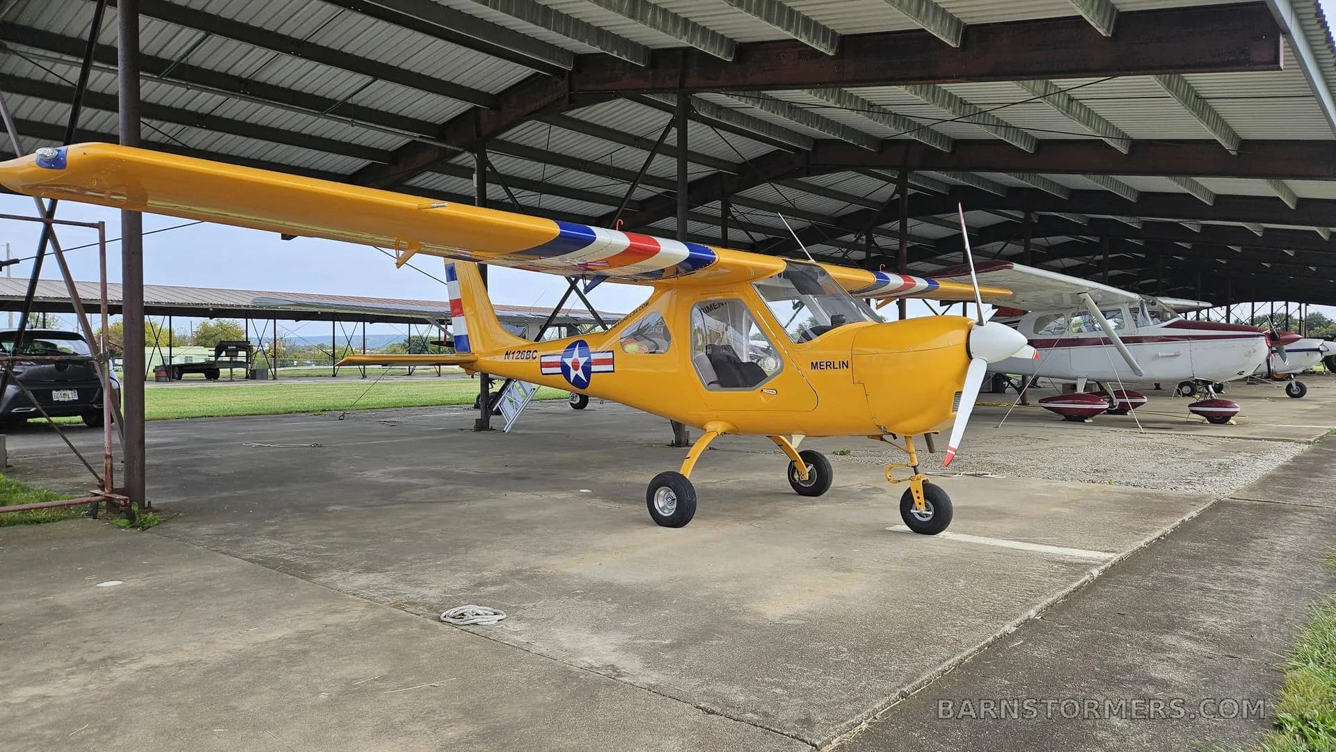 A yellow Merlin ultralight aircraft with a red, white, and blue striped wing is parked under an open-sided hangar.