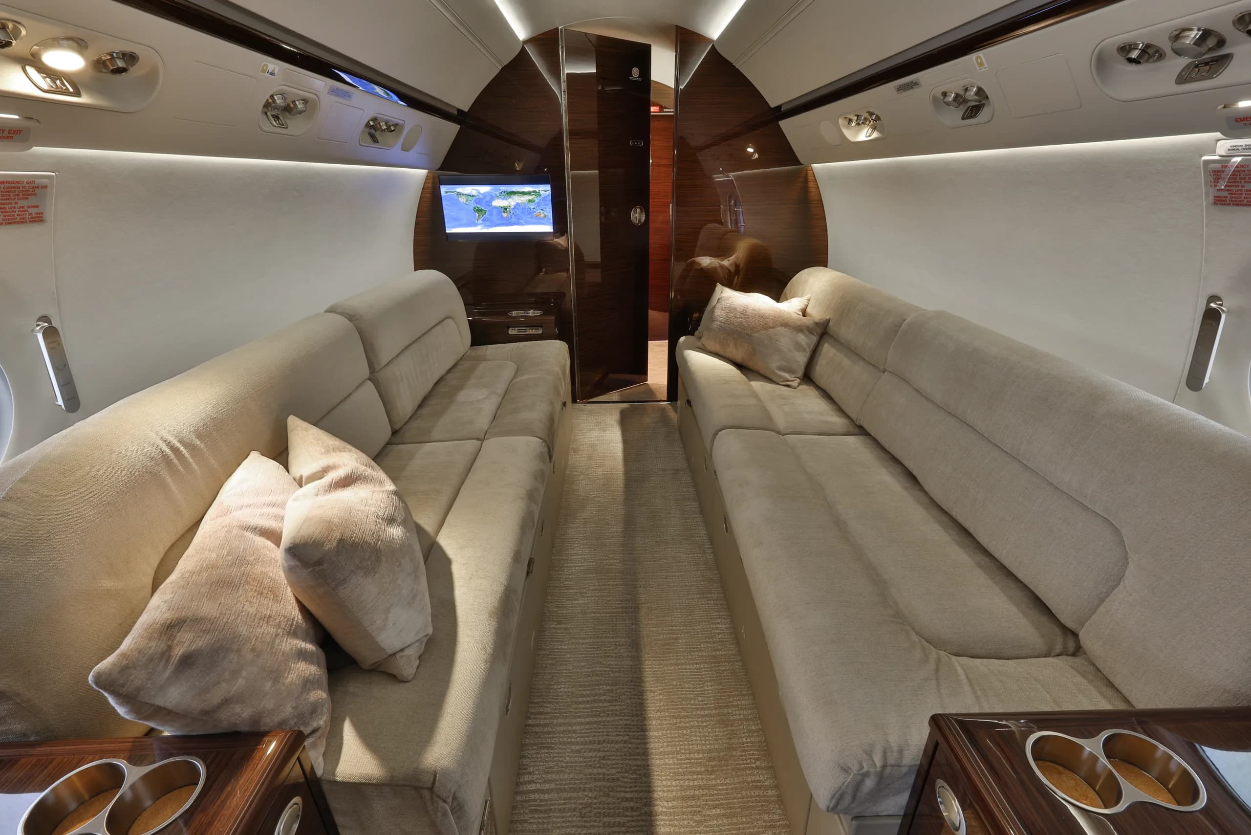 A spacious aircraft cabin featuring two long beige sofas facing each other, with a world map displayed on a screen in the background.