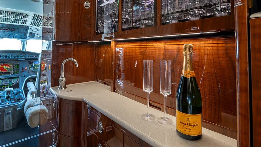 A luxurious aircraft galley with dark wood cabinetry, a sink, and a bottle of champagne with two flutes, with the cockpit visible in the background.