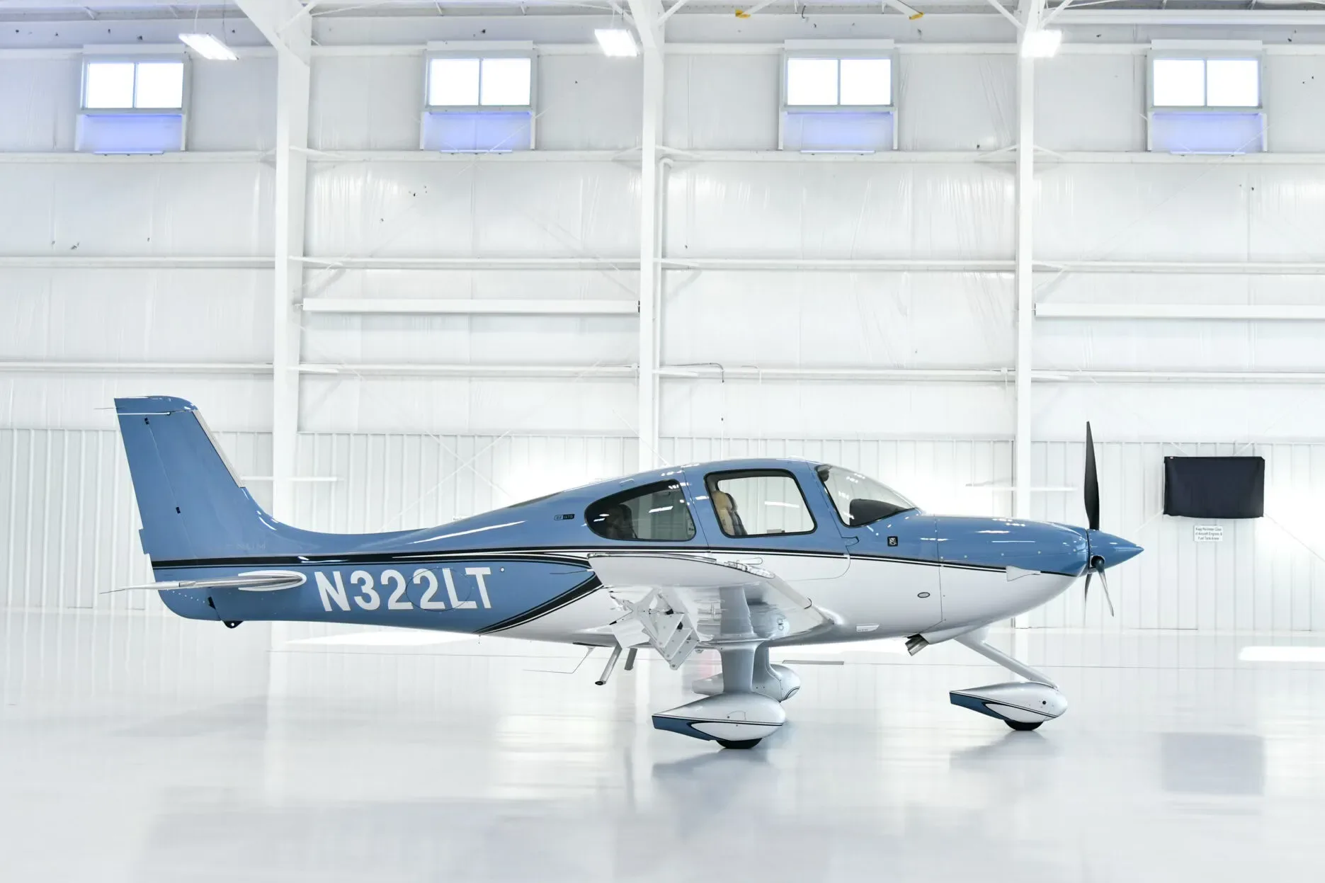 A blue and white Cirrus SR22 aircraft with registration N322LT parked in a brightly lit hangar, viewed from the side.