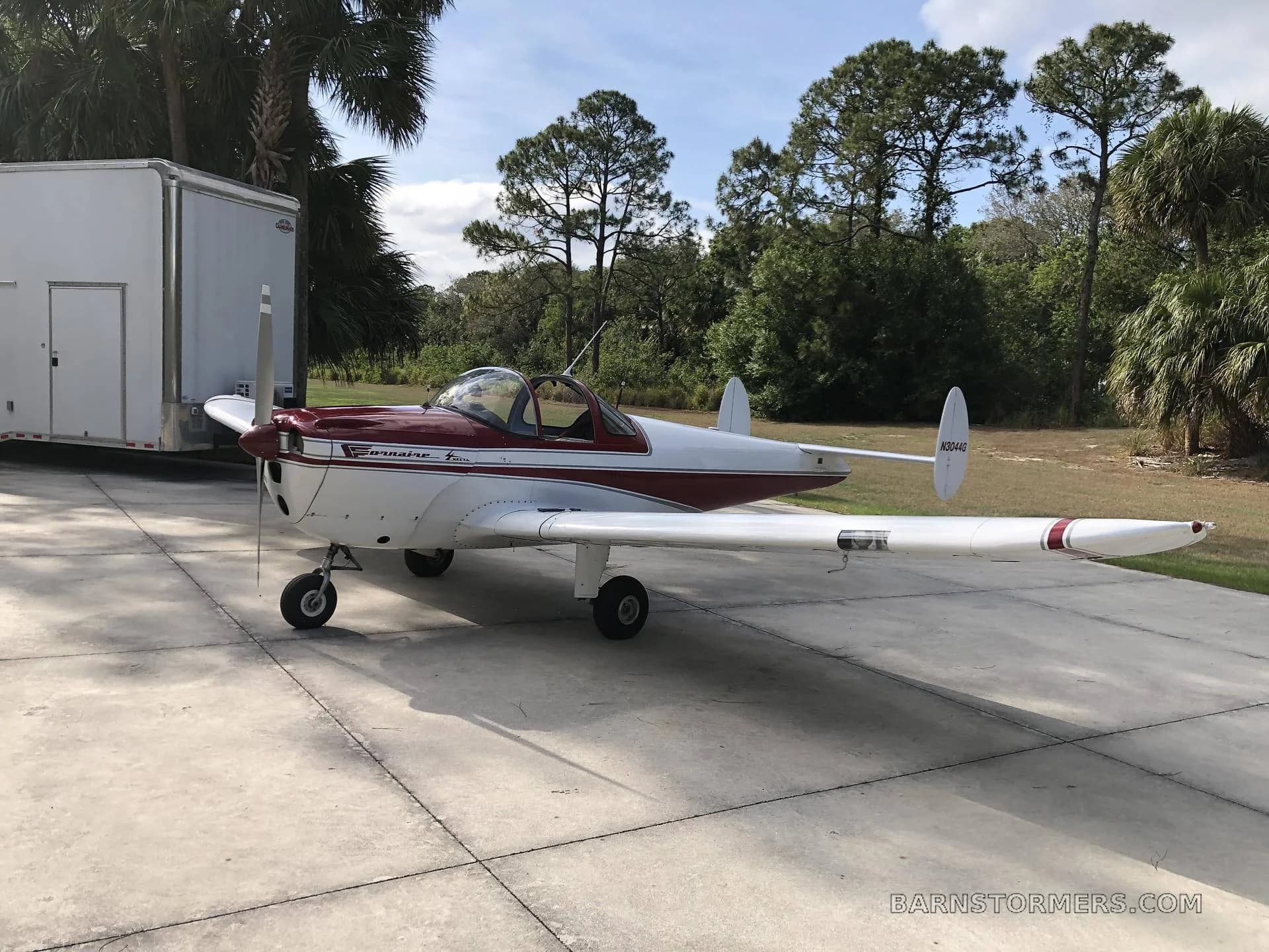A white and maroon Ercoupe 415-C aircraft with tail number N304G is parked on a concrete pad next to a white trailer and trees.