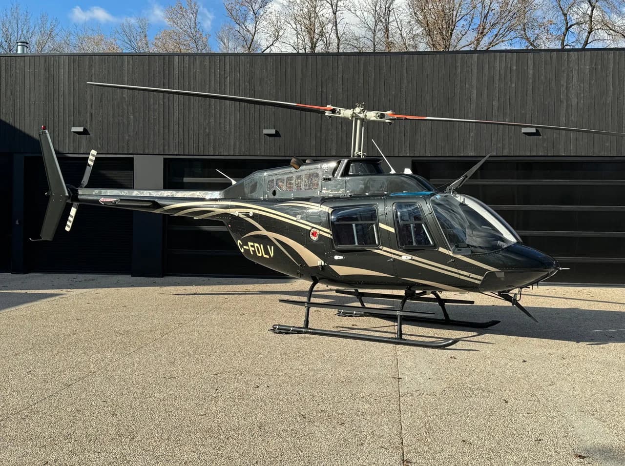 A dark green Bell 206B JetRanger III helicopter with gold stripes is parked on a concrete pad in front of a modern black building.