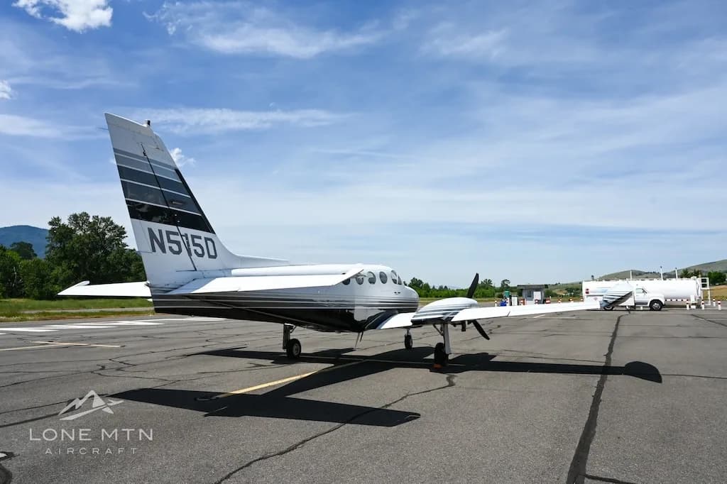 A rear-side view of the white and black twin-engine aircraft, N515D, on an airfield with a fuel truck in the distance.