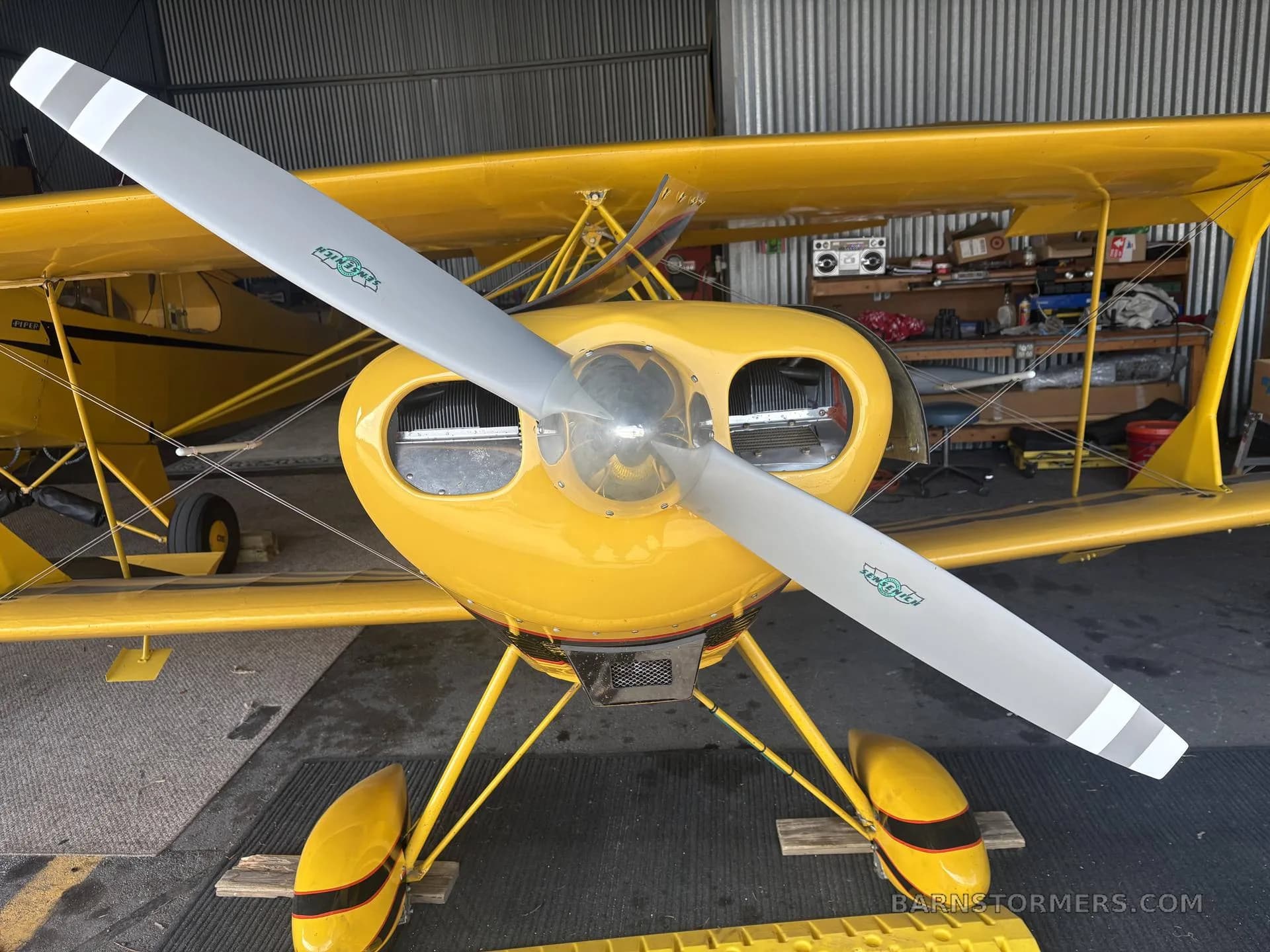 A close-up front view of a yellow biplane's propeller and engine cowling, with the aircraft parked inside a hangar.