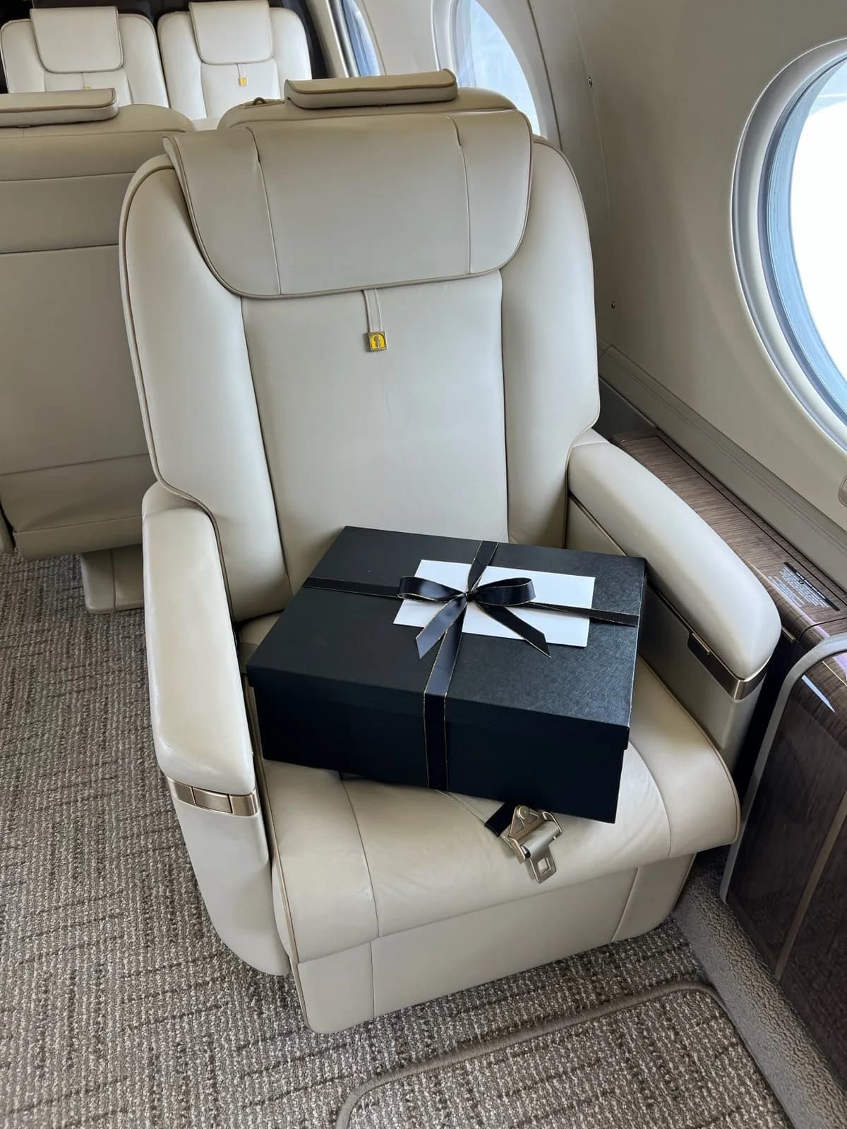 A close-up of a light-colored leather cabin seat with a black gift box tied with a ribbon resting on it.