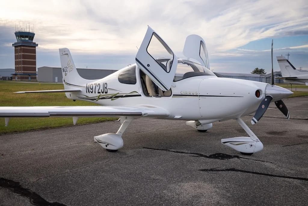 A white Cirrus SR22T aircraft, N972JB, is parked on an airfield with its doors open, an air traffic control tower in the background.