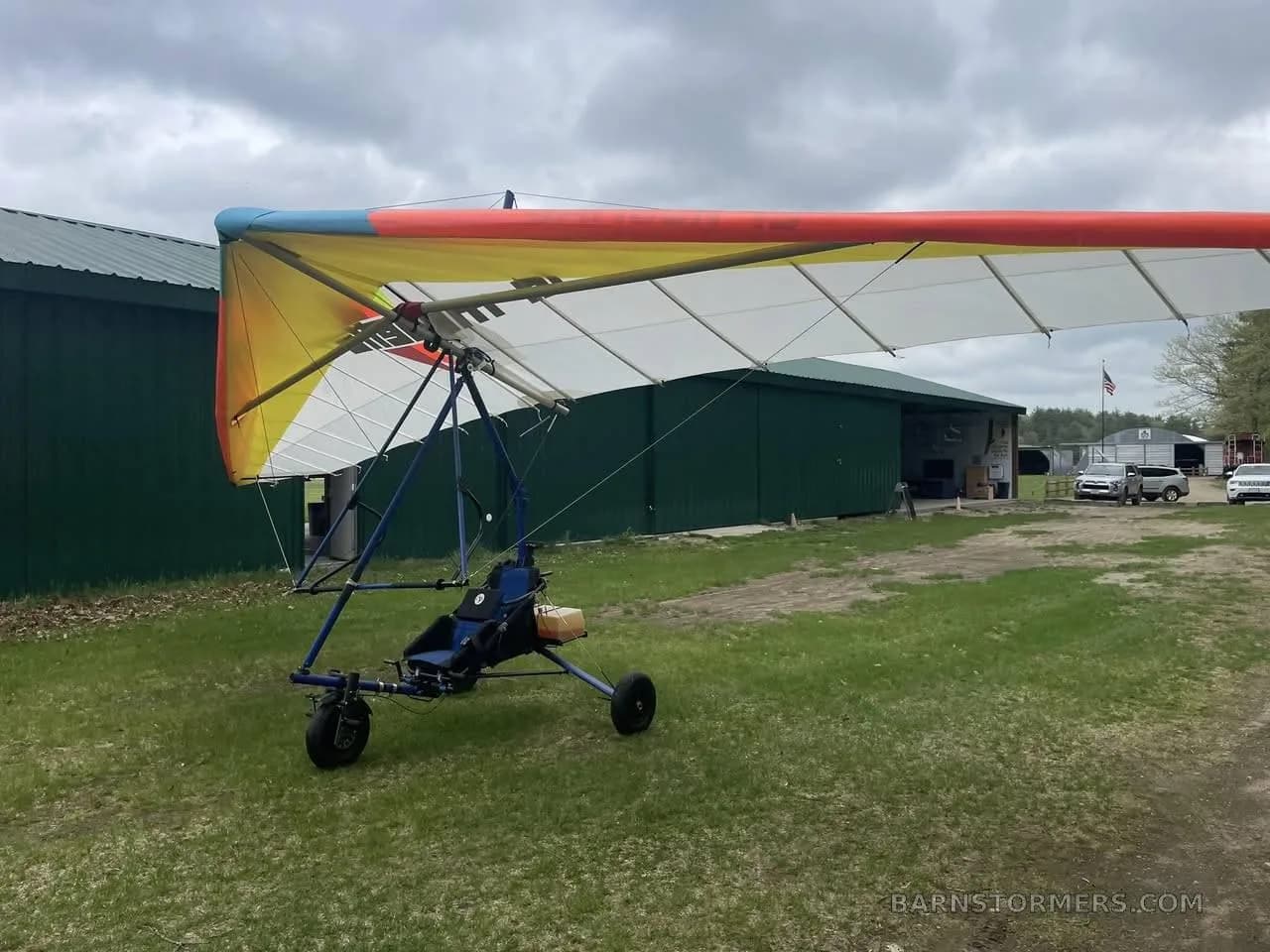 A powered hang glider with an orange, yellow, and white wing is parked on a grassy area next to a green hangar.