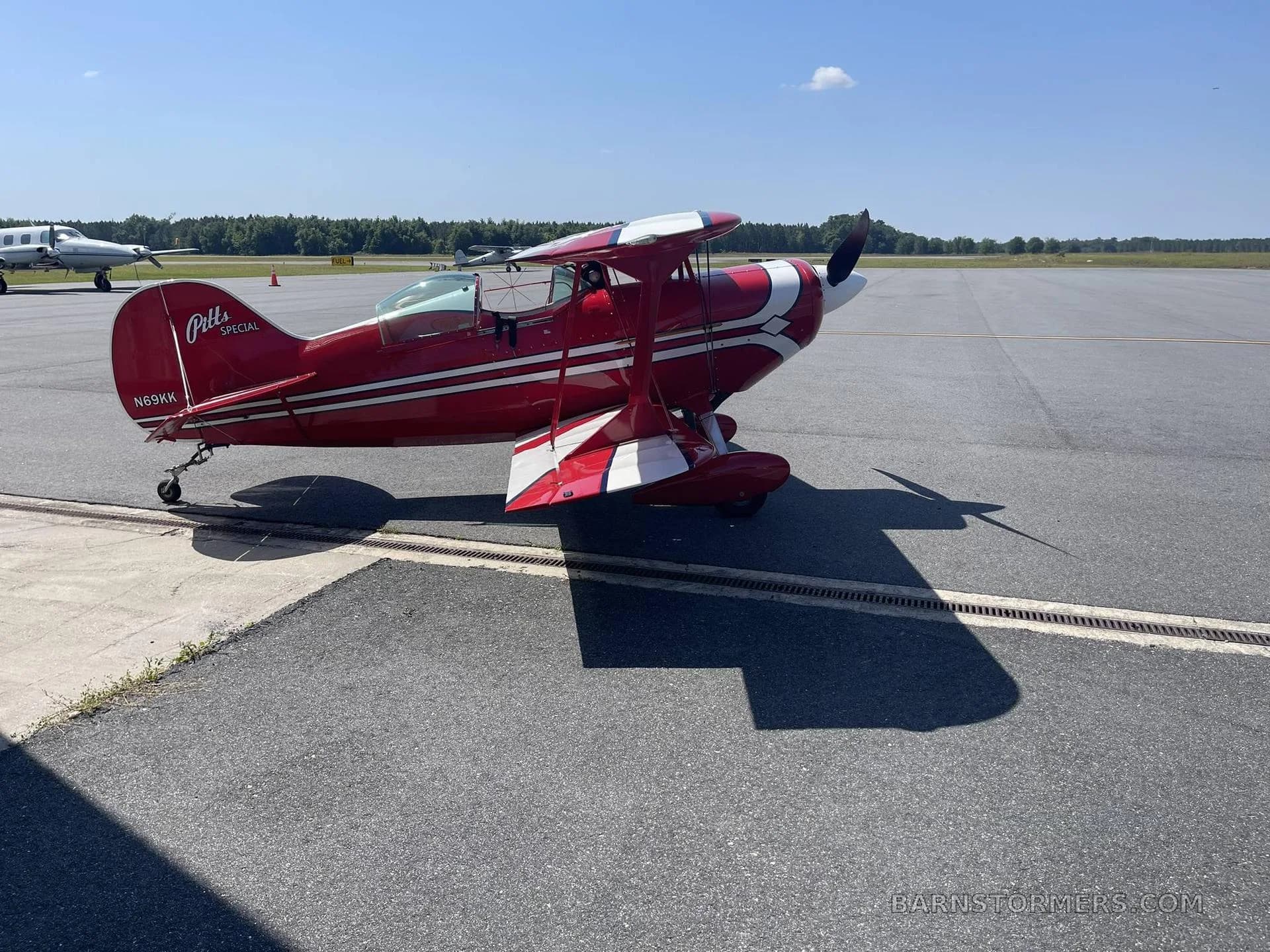 A red and white Pitts Special biplane, registration N69KK, is parked on an airfield tarmac under a clear blue sky.