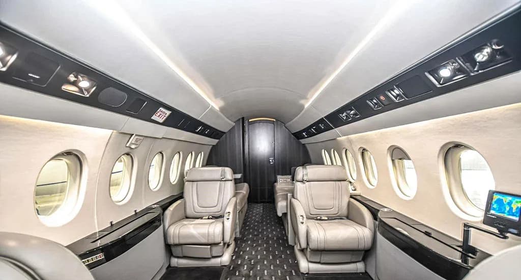 The elegant interior cabin of the Dassault Falcon 900EX, looking towards the rear with light-colored seats and dark wood accents.