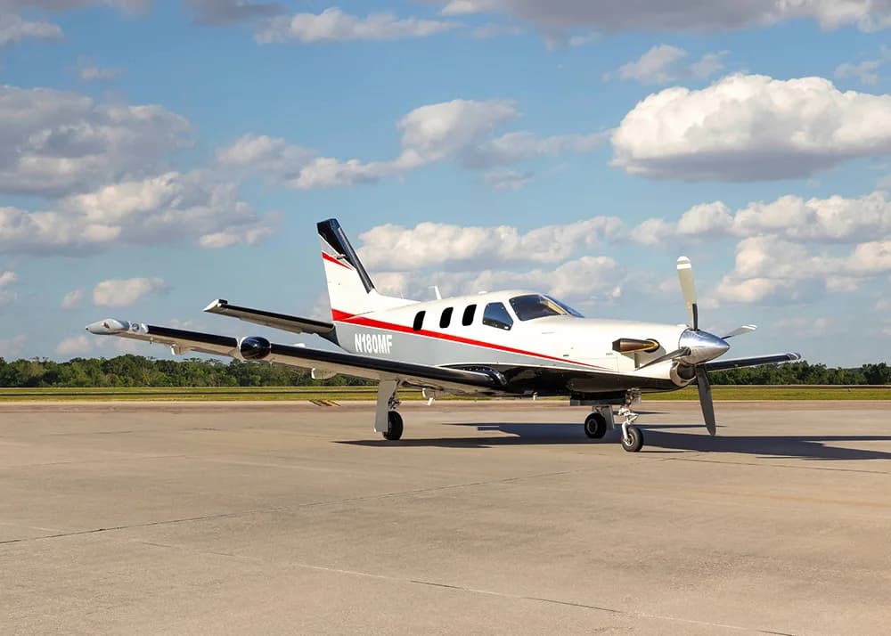 A white and grey TBM 900 series aircraft with red stripes, registration N180MF, parked on an airfield under a partly cloudy sky.