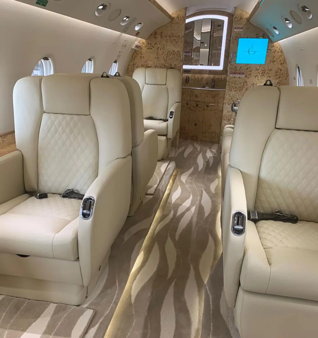 The private jet cabin interior with multiple cream leather seats arranged facing each other, and a patterned aisle carpet.