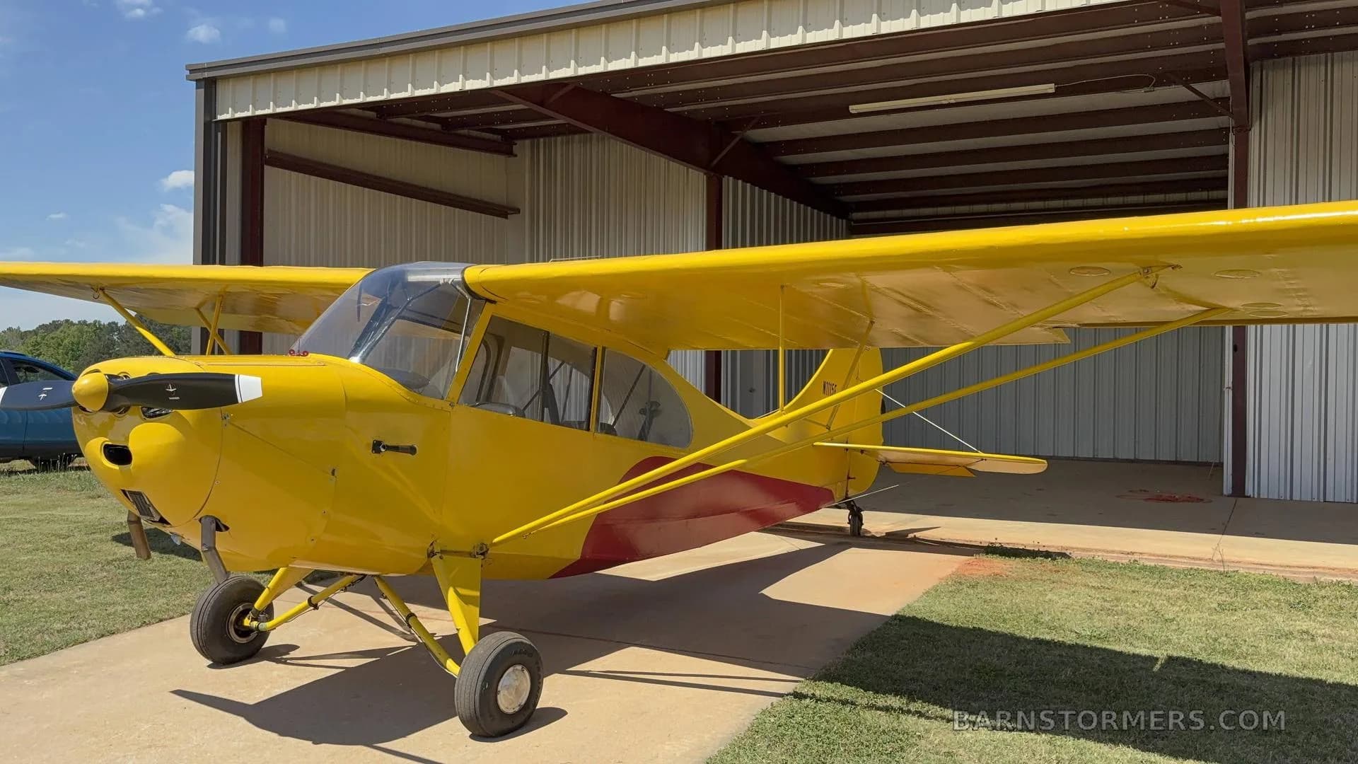 A yellow single-engine aircraft with red accents, parked on a concrete pad outside a hangar.
